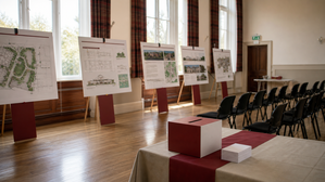 Community hall set up for a public consultation event, with plan display boards, a feedback box and rows of empty chairs, lit by daylight through tall windows.