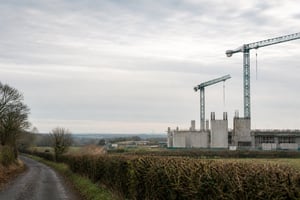 A UK infrastructure construction site under an overcast sky, with tower cranes on the right and a quiet rural lane in the foreground.